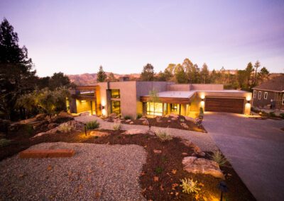 Modern house with illuminated windows at twilight, featuring a landscaped garden and a concrete driveway.