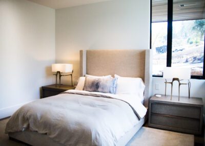 Modern bedroom with a neatly made bed, two bedside tables, and lamps, featuring large windows with a view outside.