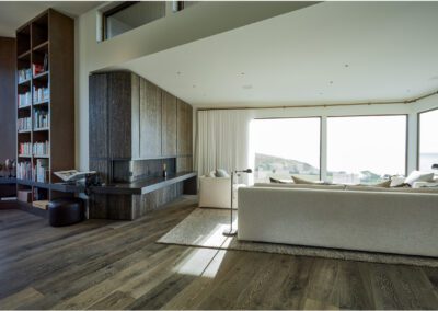 Modern living room with floor-to-ceiling windows offering a coastal view, minimalist furniture, and wooden flooring.