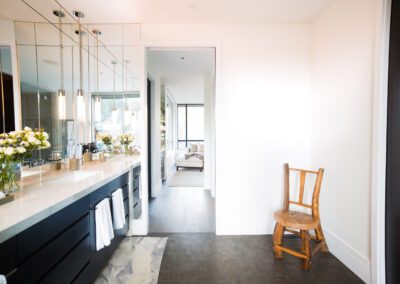 A bright, modern bathroom with double vanity and a wooden chair, leading to an adjoining room.
