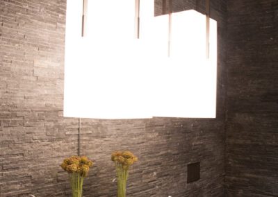 Modern bathroom with sleek design, featuring a large rectangular light fixture, textured dark walls, and decorative towels and plants on the vanity.